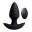 A black vibrating silicone butt plug with a tapered tip stands next to its remote control. 